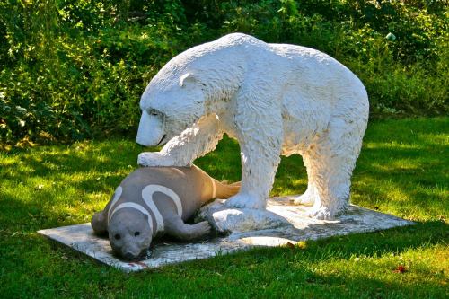 A white bear sculpture stands over a brown seal sculpture on a grassy area, surrounded by trees.