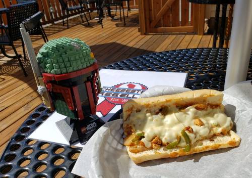 A LEGO Boba Fett figure next to a cheesy chicken sandwich on a table with a logo in the background.