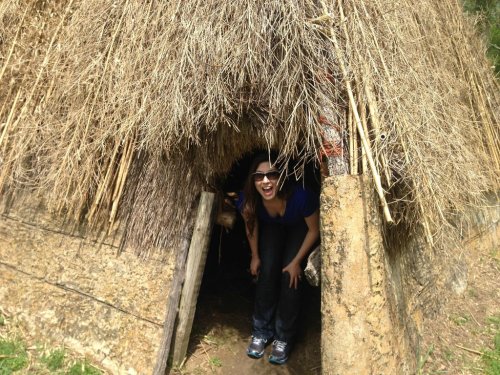 A person smiling while standing in the entrance of a thatched hut surrounded by greenery.