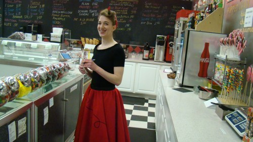 A smiling person in a black top and red skirt stands behind a counter filled with candy and snacks.
