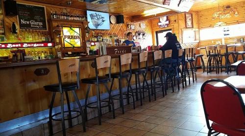 A cozy bar interior with wooden decor, empty stools, and a bartender serving drinks. A patron sits at the counter.
