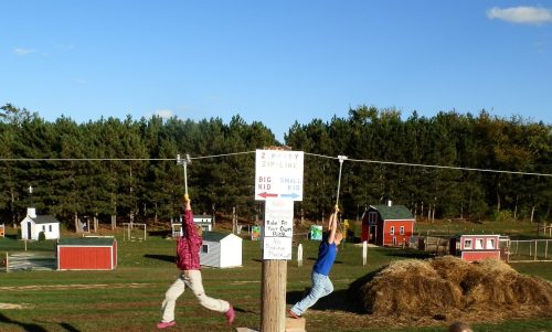 Two children ziplining on a sunny day, with a sign indicating "Big Kid" and "Small Kid" zipline options in the background.