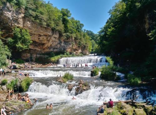 A scenic waterfall surrounded by lush greenery, with people enjoying the water and sun on a clear day.