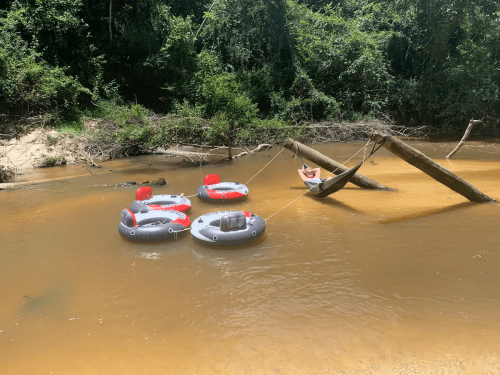 Four inflatable rafts float in a muddy river, with a hammock tied between trees nearby. Lush greenery surrounds the scene.