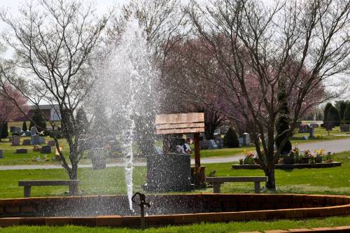 A fountain sprays water in a cemetery, surrounded by trees and flower beds, with gravestones in the background.