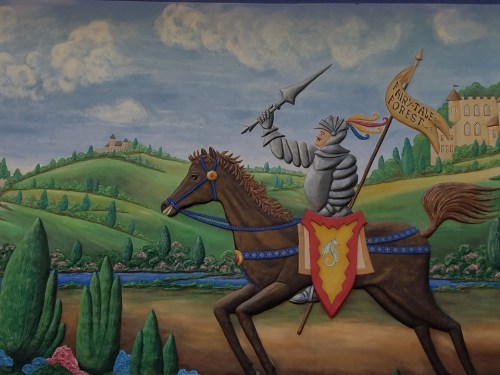 A knight in armor rides a horse, holding a flag, with a colorful landscape of hills and flowers in the background.