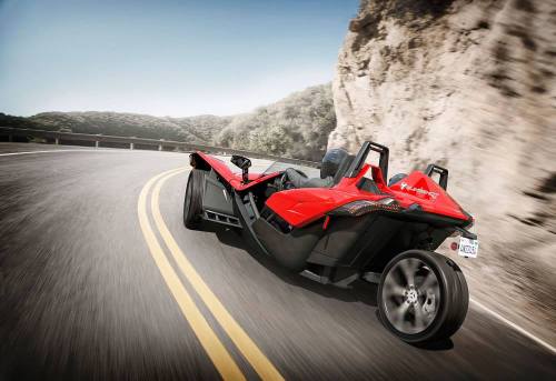 A red three-wheeled sports car driving on a winding road with a mountainous backdrop.