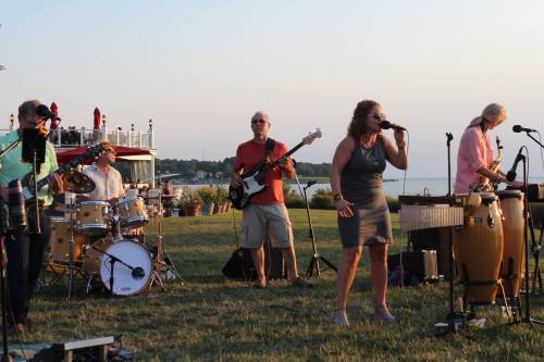 A band performs outdoors at sunset, featuring a singer, guitarists, and a drummer, with a scenic waterfront backdrop.