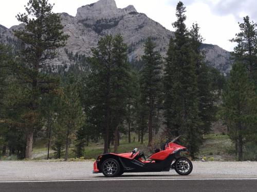 A red three-wheeled vehicle parked on a road, surrounded by tall pine trees and mountains in the background.