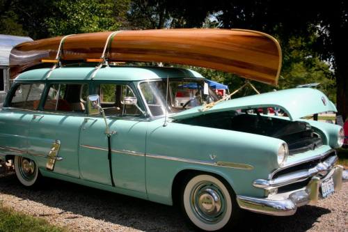 A vintage turquoise car with a wooden canoe on its roof, parked on a gravel surface.