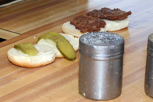Two burger patties on buns with pickles, alongside a metal salt shaker on a wooden countertop.