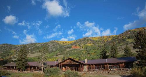 A rustic lodge surrounded by trees and mountains under a blue sky with scattered clouds.