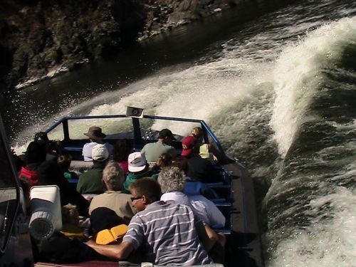 A group of people on a boat, splashing through water with waves and rocky shores in the background.
