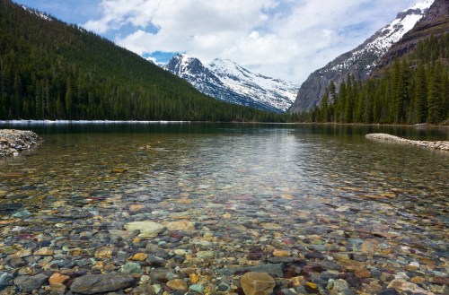A serene lake with clear water reflecting mountains and lush green trees under a partly cloudy sky.
