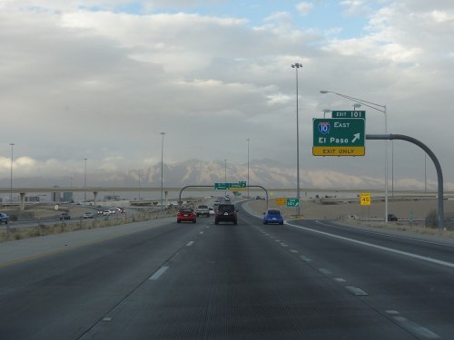 Highway scene with vehicles, directional signs for El Paso, and mountains in the background under a cloudy sky.