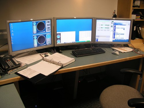 A workspace with three computer monitors displaying data, a keyboard, a phone, and a binder on a desk.
