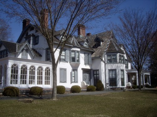 A large, elegant white house with multiple gables, large windows, and a manicured lawn under a clear blue sky.
