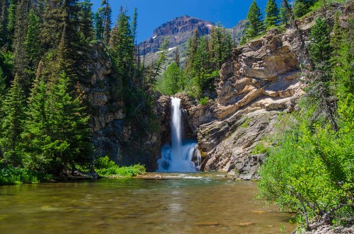 A serene waterfall cascades down rocky cliffs, surrounded by lush greenery and tall trees under a clear blue sky.