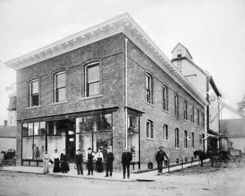 Historic black-and-white photo of a brick building with people standing outside, including men and women in period clothing.