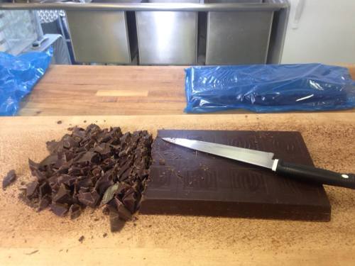 A large block of chocolate is partially chopped on a wooden surface, with a knife resting beside it.