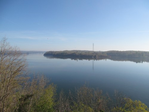 A serene lake reflects the clear blue sky, surrounded by trees and distant hills, with a power line tower in view.