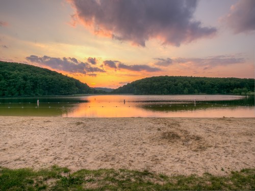 Sunset over a tranquil lake, surrounded by lush hills, with a sandy beach in the foreground and clouds in the sky.