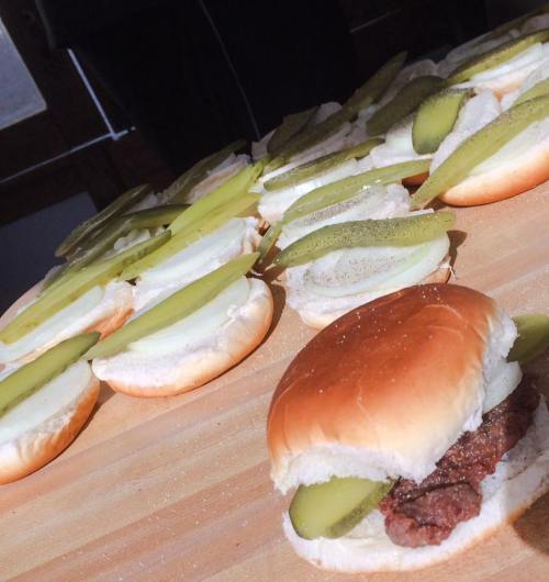A close-up of a burger with pickles on a wooden surface, with several buns and pickles in the background.