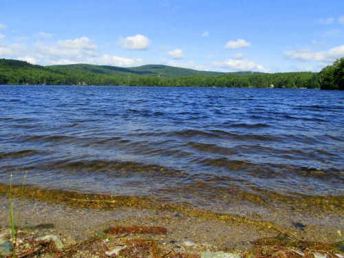 A serene lake with gentle waves, surrounded by lush green hills and a clear blue sky dotted with clouds.