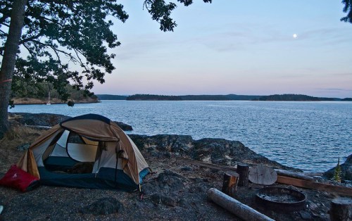 A tent by the water at dusk, with a calm lake and distant trees under a moonlit sky.