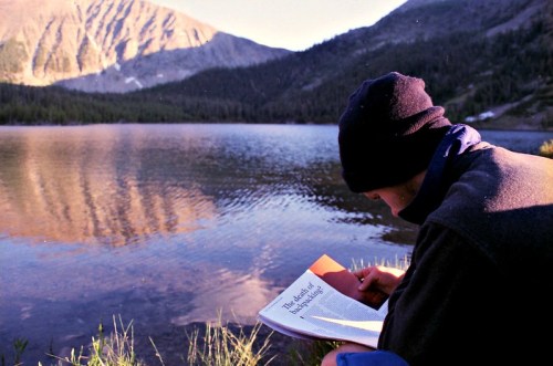 A person reading by a serene lake, surrounded by mountains and trees, with sunlight reflecting on the water.