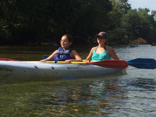 Two people kayaking on a calm river, one wearing a life jacket and the other in a tank top, enjoying a sunny day.