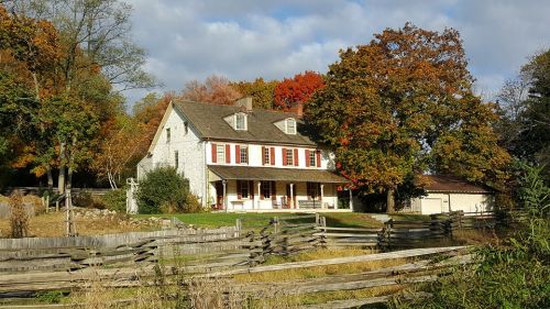 A charming white house with red shutters surrounded by colorful autumn trees and a wooden fence.