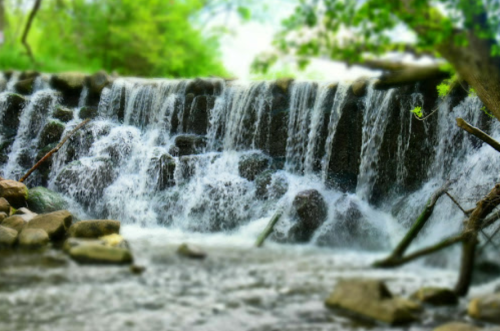 A serene waterfall cascading over rocks, surrounded by lush greenery and a gentle stream below.
