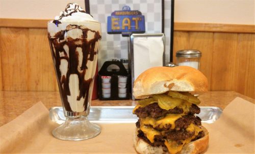 A delicious cheeseburger with pickles next to a tall glass of chocolate sundae topped with whipped cream.