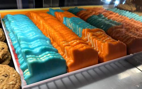 Colorful layered desserts in blue and orange, arranged neatly in a display case, with cookies visible on the side.
