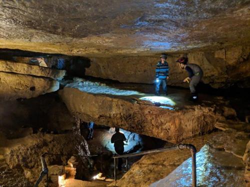 Two people explore a dimly lit cave, using flashlights to illuminate the rocky terrain and formations around them.