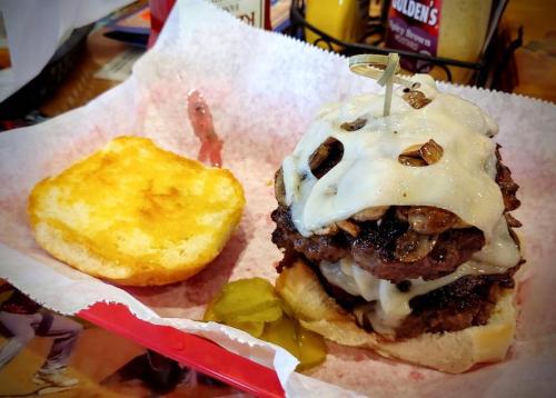 A stacked burger with melted cheese, mushrooms, and a toothpick, served with a toasted bun and pickles on the side.