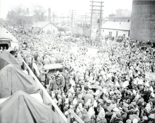 A large crowd gathers near train tracks, holding signs, with buildings and power lines in the background.