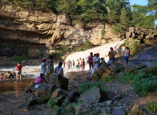People enjoying a sunny day by a waterfall, surrounded by rocky terrain and lush greenery.