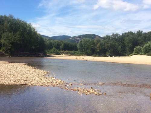 A serene river scene with sandy banks, surrounded by trees and mountains under a blue sky with scattered clouds.