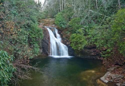 A serene waterfall cascades into a calm pool, surrounded by lush greenery and rocky terrain.