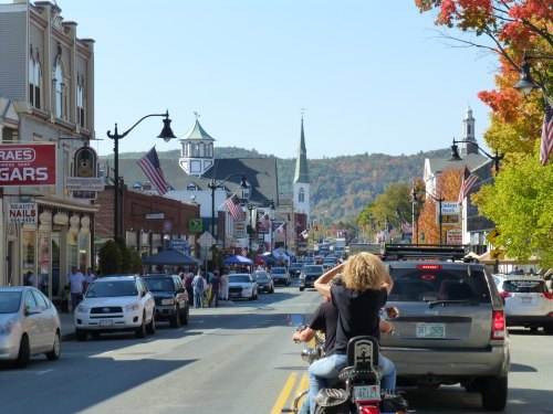 A bustling small town street lined with shops, flags, and colorful autumn trees, with a church steeple in the distance.