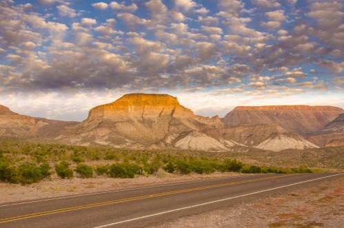 A scenic view of a desert landscape with a mountain plateau under a cloudy sky, alongside a winding road.