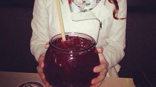 A person holding a large fishbowl drink with colorful straws, sitting at a table in a restaurant.