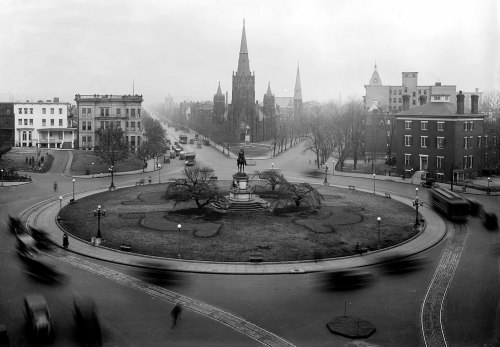 Historic black-and-white photo of a city square with a statue, churches, and early 20th-century vehicles.