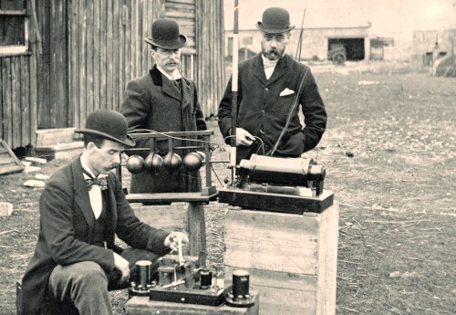 Three men in bowler hats examine early electrical equipment outdoors, with wooden structures in the background.