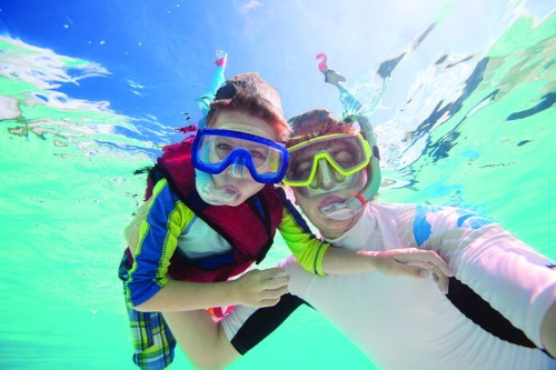 A child and adult wearing snorkeling gear underwater, smiling and enjoying a sunny day in clear blue water.