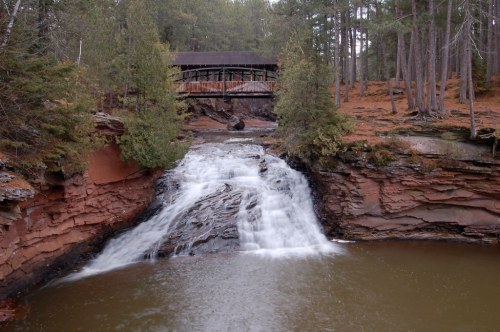 A serene waterfall cascades over rocky terrain, with a wooden bridge in the background surrounded by tall trees.