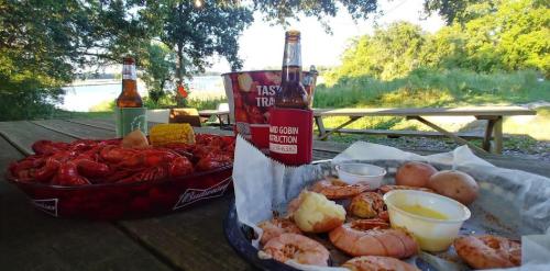 A picnic table with a spread of crawfish, shrimp, potatoes, and drinks, set by a scenic lakeside view.