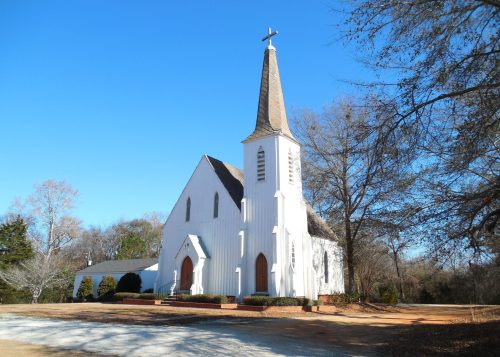 A white church with a tall steeple and cross, surrounded by trees under a clear blue sky.
